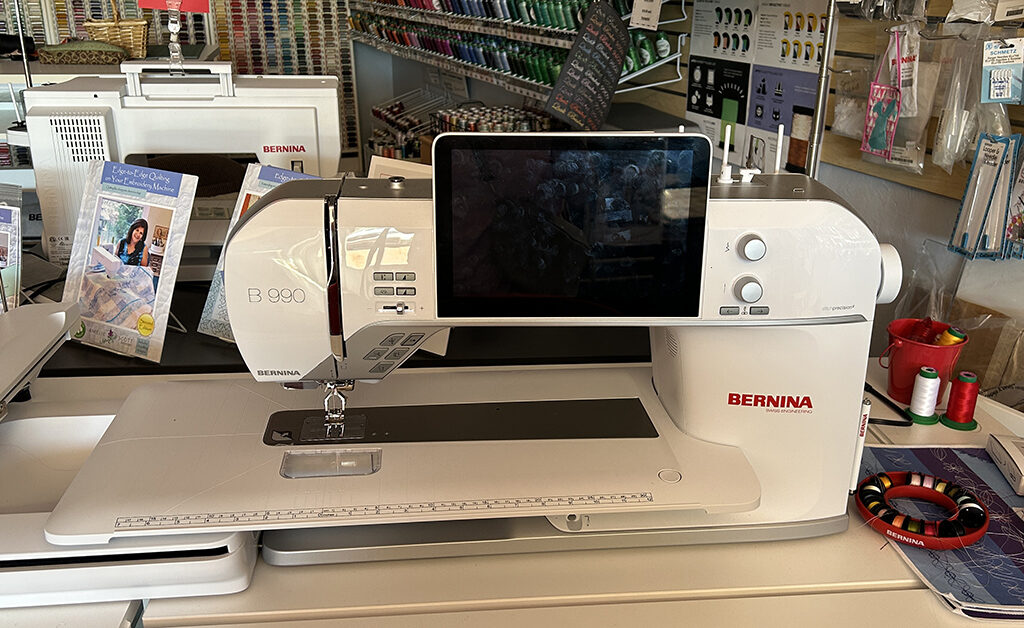 A big white Bernina B 990 sewing machine with a large touchscreen sits on a table in a sewing shop. There are spools of thread, sewing tools, and quilting books around it.